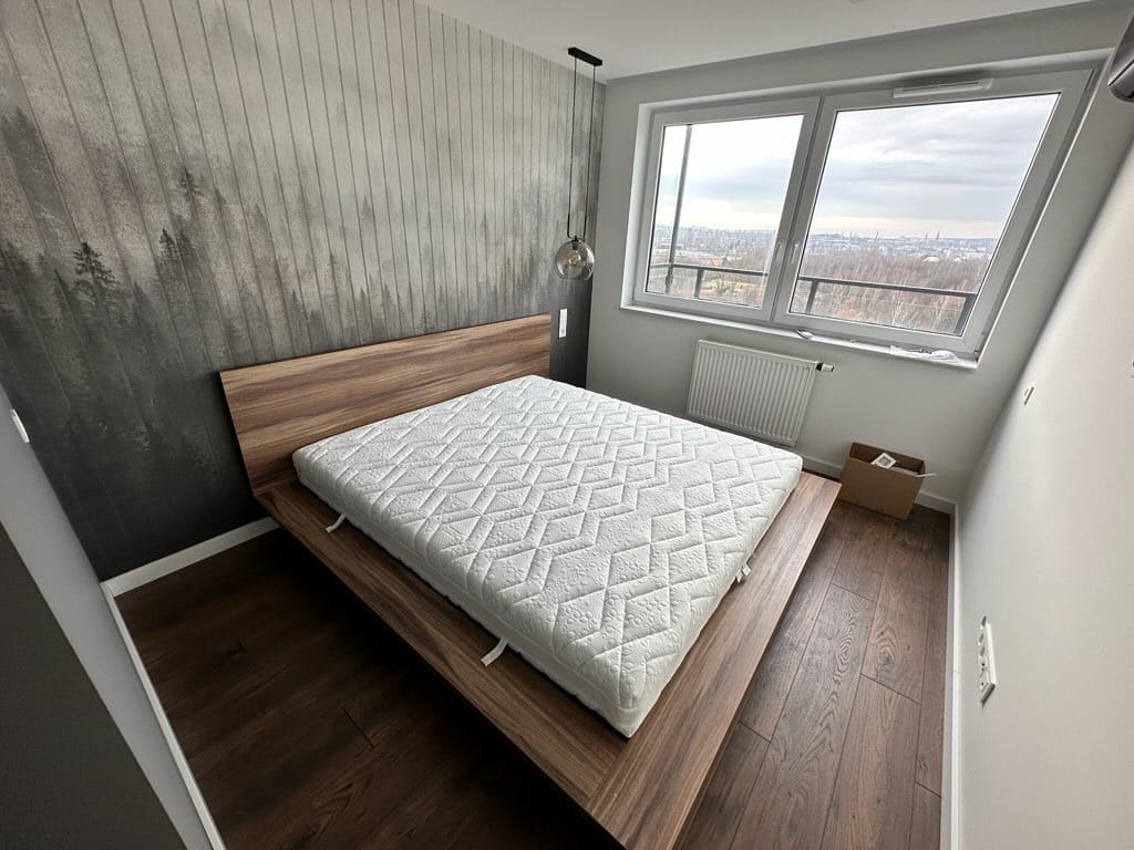 Nowoczesna sypialnia z drewnianą ramą łóżka i tapetą z motywem lasu, widok na miasto z okna