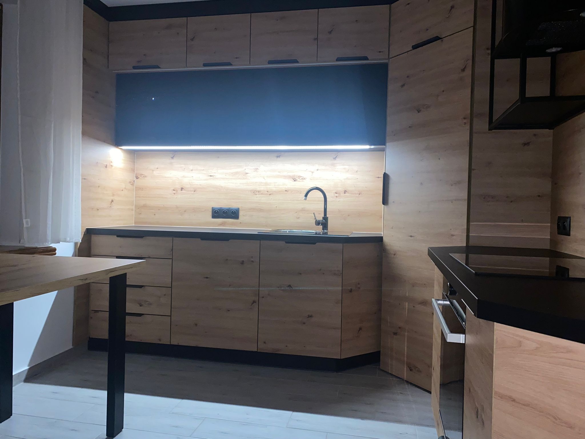 Nowoczesna kuchnia z drewnianymi frontami, czarnym blatem i oświetleniem LED pod szafkami wiszącymi, widoczny fragment stołu z czarnymi nogami.
