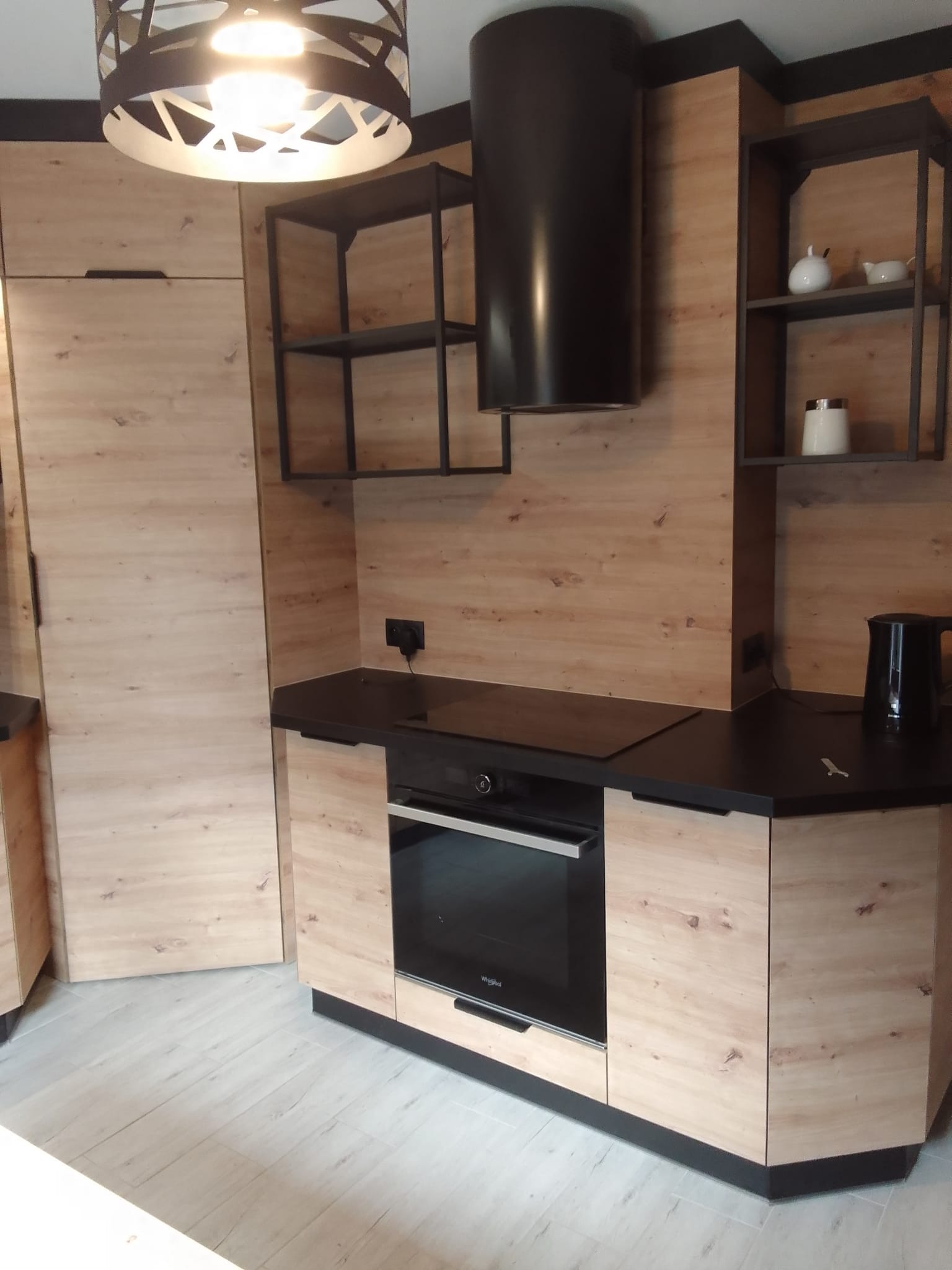 Nowoczesna kuchnia z frontami w odcieniach jasnego drewna, czarnym blatem i czarnymi akcentami w postaci okapu, półek i oświetlenia, minimalistyczny design.