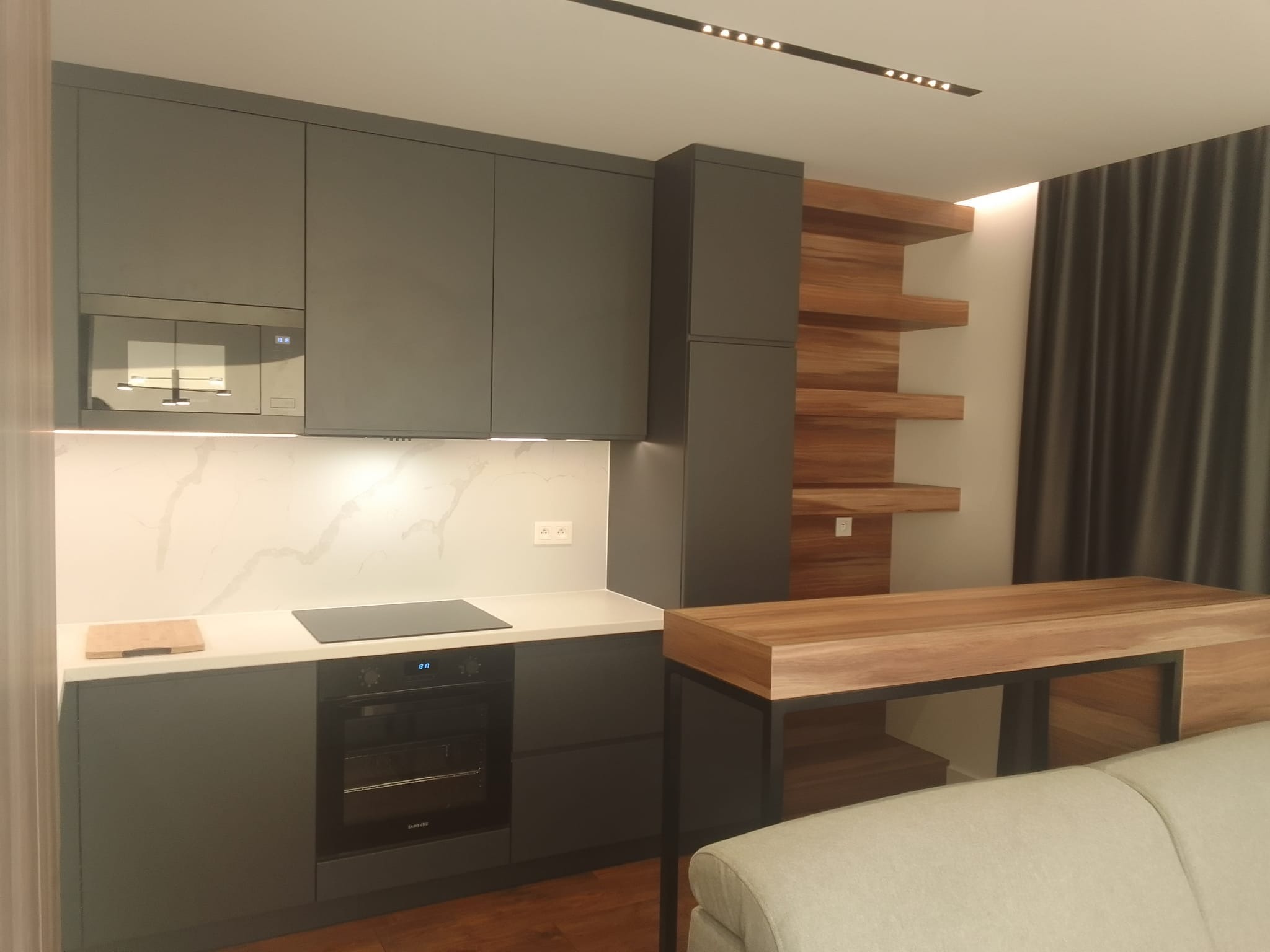 Nowoczesna kuchnia w odcieniach szarości z drewnianymi akcentami, minimalistyczne szafki, biały blat z marmurowym wzorem i indukcyjną płytą grzewczą, drewniane półki na ścianie, oświetlenie...