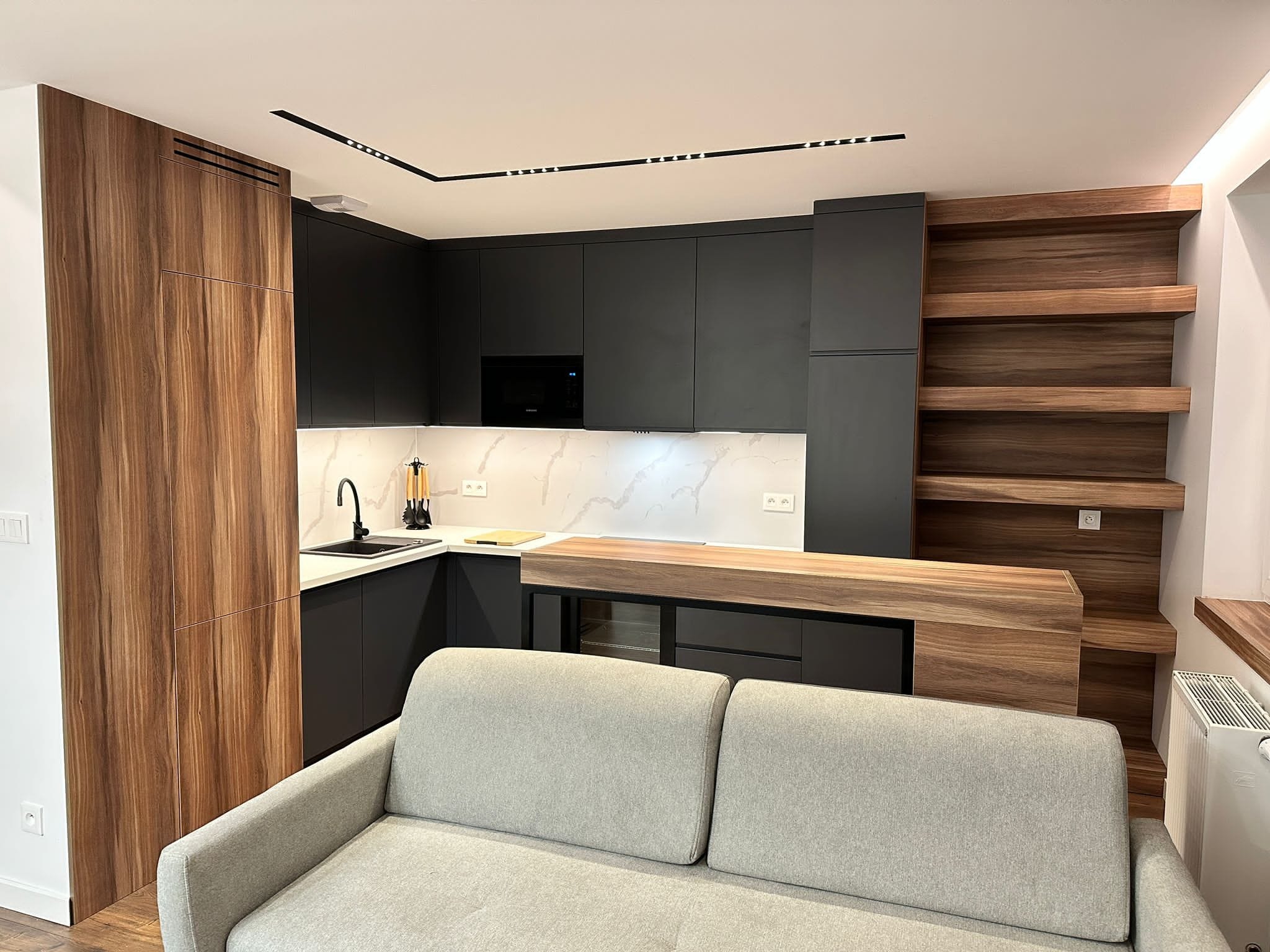 Nowoczesna kuchnia z czarnymi szafkami i blatem imitującym marmur, połączona z drewnianą zabudową i półkami, widoczna zza szarej sofy.
