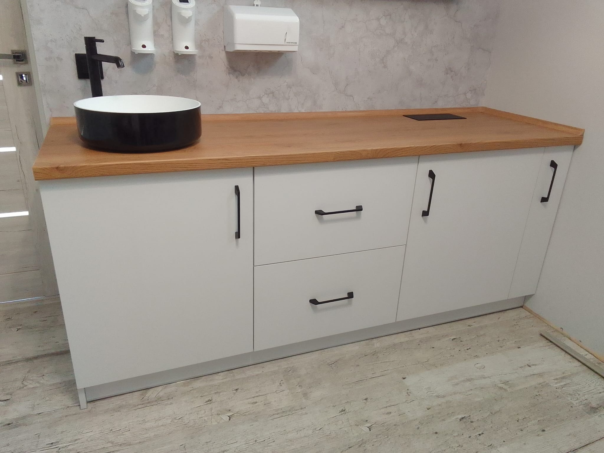 Białe szafki łazienkowe z drewnianym blatem, czarną umywalką nablatową i baterią, oraz dozowniki mydła na ścianie o wzorze przypominającym beton.