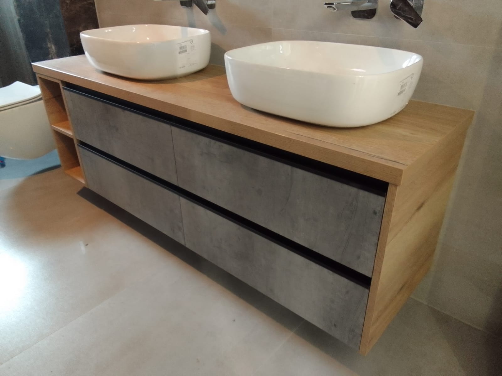 Nowoczesna szafka łazienkowa z dwiema umywalkami nablatowymi, drewnianym blatem i szarymi frontami szuflad z czarnymi akcentami, widoczna toaleta w tle po lewej stronie.