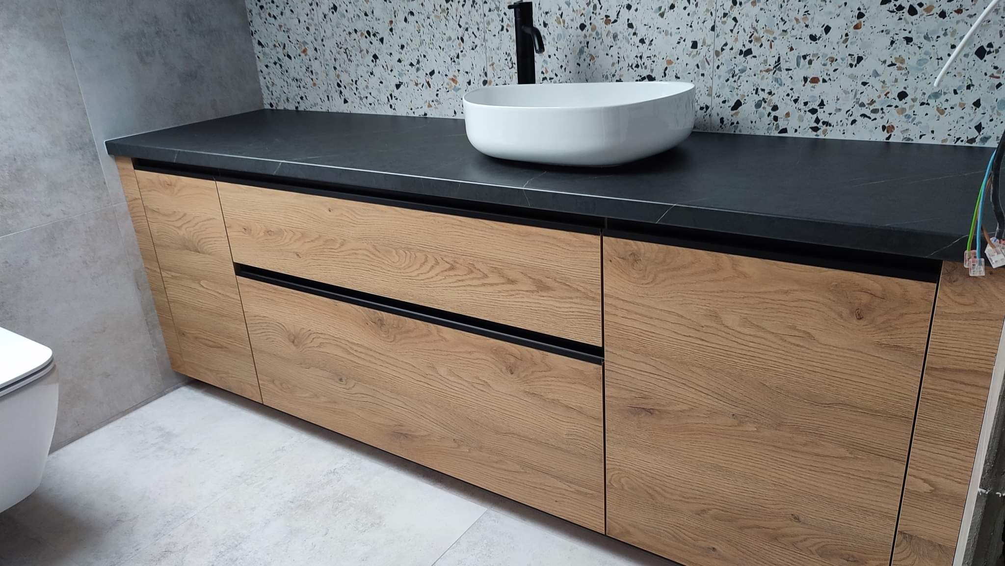 Łazienka z drewnianą szafką pod umywalkę z czarnym blatem i owalną białą umywalką nablatową, na ścianie mozaika.
