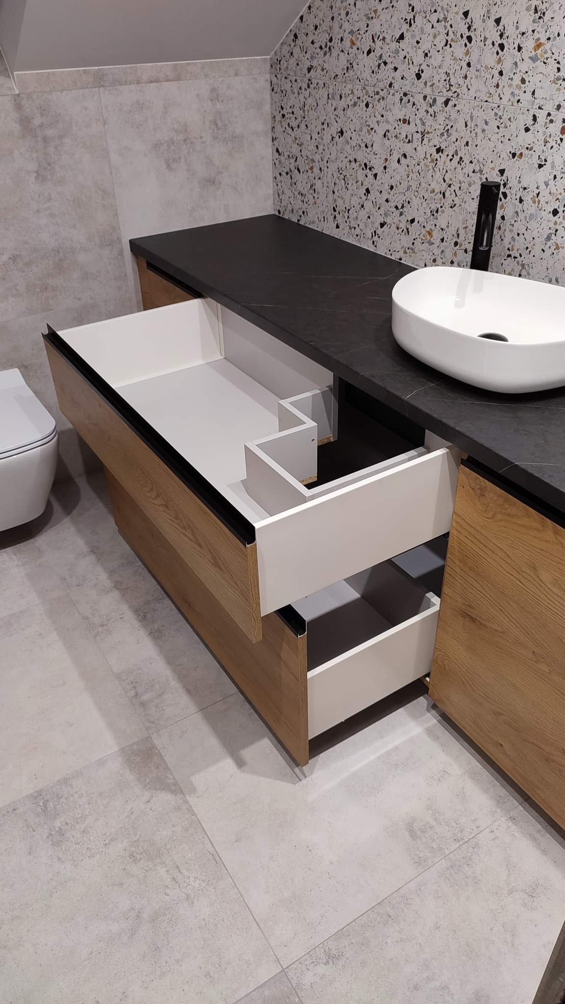 Łazienkowa szafka pod umywalkę z otwartymi szufladami o drewnianym froncie i białym wnętrzu, czarny blat, biała umywalka nablatowa, ściana wykończona płytkami w stylu lastryko.