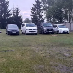 Cztery zaparkowane samochody osobowe i busy różnych marek (Seat, Volkswagen, Ford) na trawiastym terenie, z widocznym fragmentem ogrodzenia i drzew w tle.