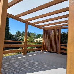 AS DOM Arkadiusz Stępka - Drewniana pergola z zadaszeniem i ażurowymi ściankami, widok z zewnątrz, jasne drewno, szara podłoga, błękitne niebo w tle, nowoczesny design.
