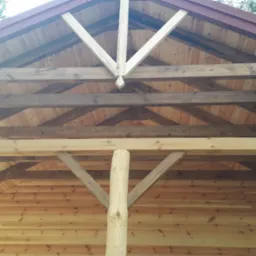 Drewniana konstrukcja dachu altany z widoczną więźbą, krokwiami i słupami nośnymi, pokryta czerwoną blachodachówką.