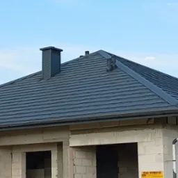 Nowy, ciemnoszary dach z kominem i orynnowaniem na surowym budynku z białego pustaka, widok z dołu.