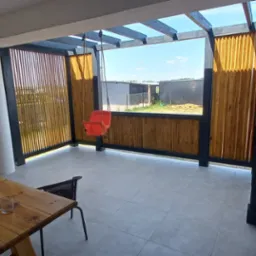 Zadaszony taras z drewnianymi panelami, czarną konstrukcją i huśtawką dla dziecka, widok na podwórze z budynkami gospodarczymi.