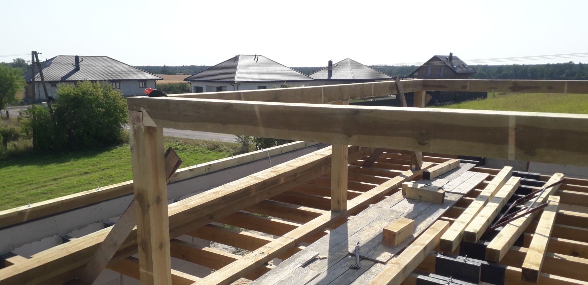 Wznoszenie drewnianej konstrukcji dachu na nowym budynku, widok z góry na elementy więźby dachowej, w tle domy jednorodzinne i zielone tereny.