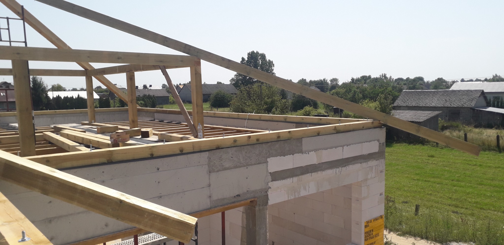 Wznoszenie drewnianej konstrukcji dachu na budynku z betonu komórkowego, widoczne elementy więźby dachowej, w tle zabudowa wiejska.
