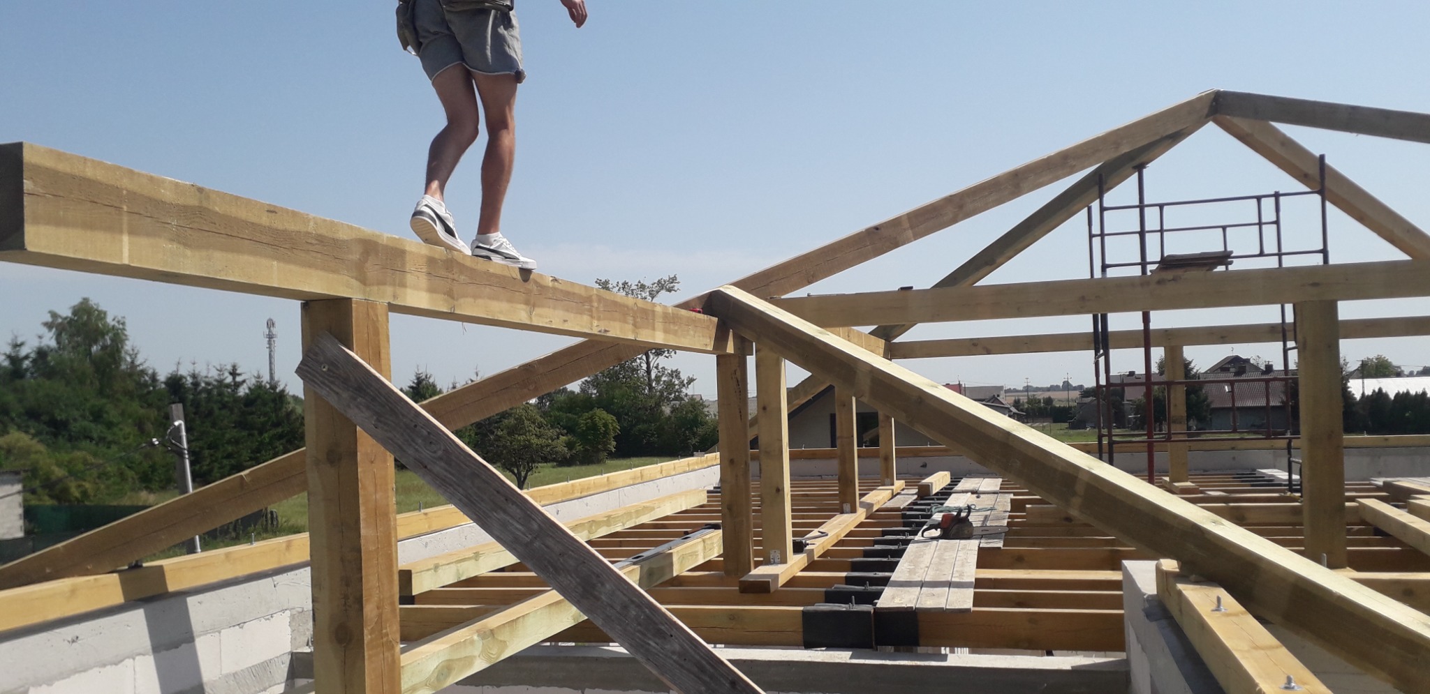Konstrukcja drewniana więźby dachowej w trakcie montażu, z pracownikiem w sportowych butach chodzącym po belce, widok na otwartą przestrzeń i okoliczne budynki w tle.