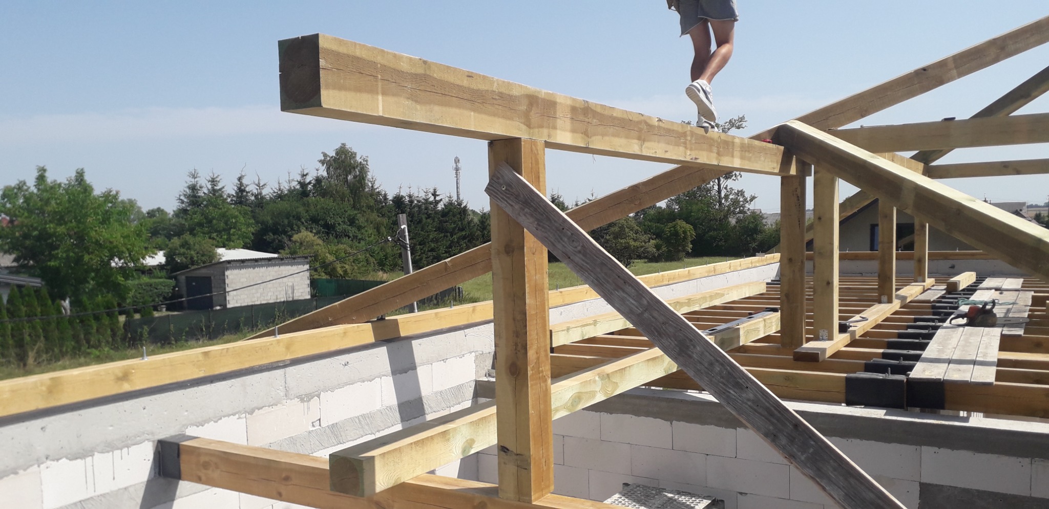 Konstrukcja drewniana więźby dachowej w trakcie montażu, widoczne elementy nośne i fragment dachu w budowie, na tle błękitnego nieba i zieleni drzew, robotnik w butach sportowych na belce.