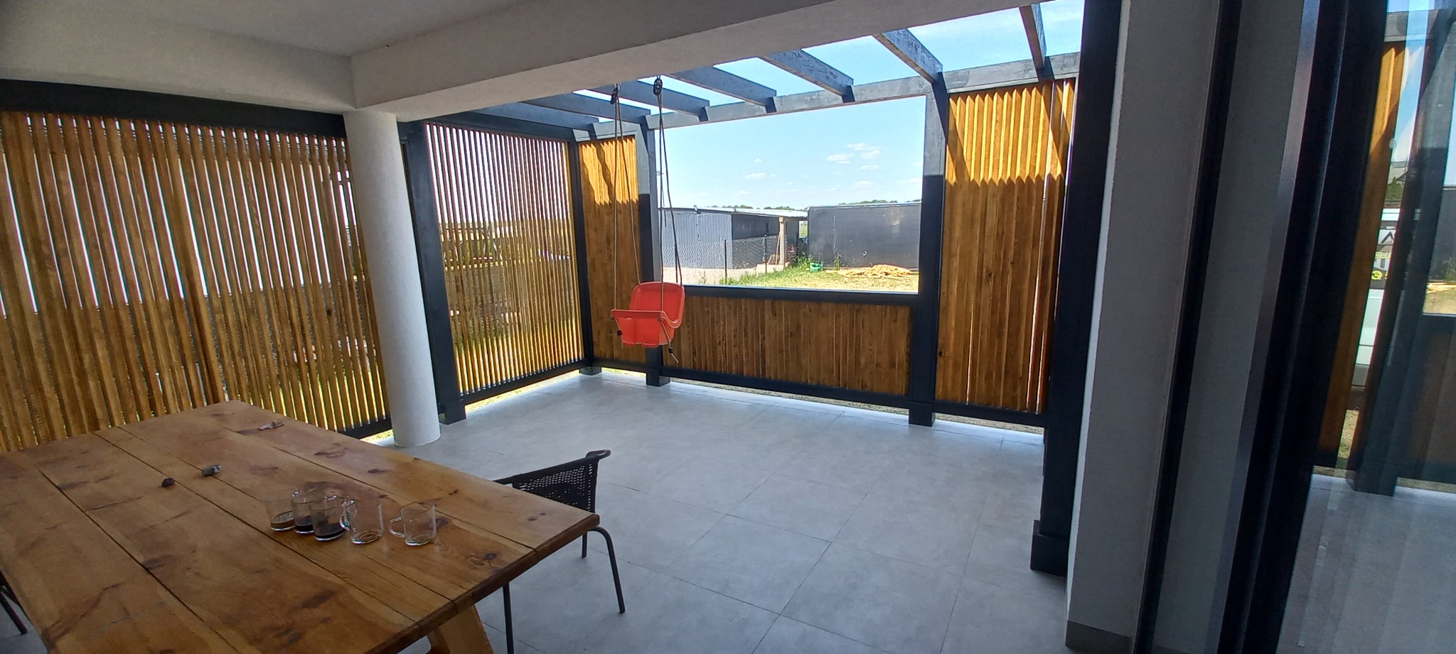 Zadaszony taras z drewnianymi panelami, czarną konstrukcją i huśtawką dla dziecka, widok na podwórze z budynkami gospodarczymi.