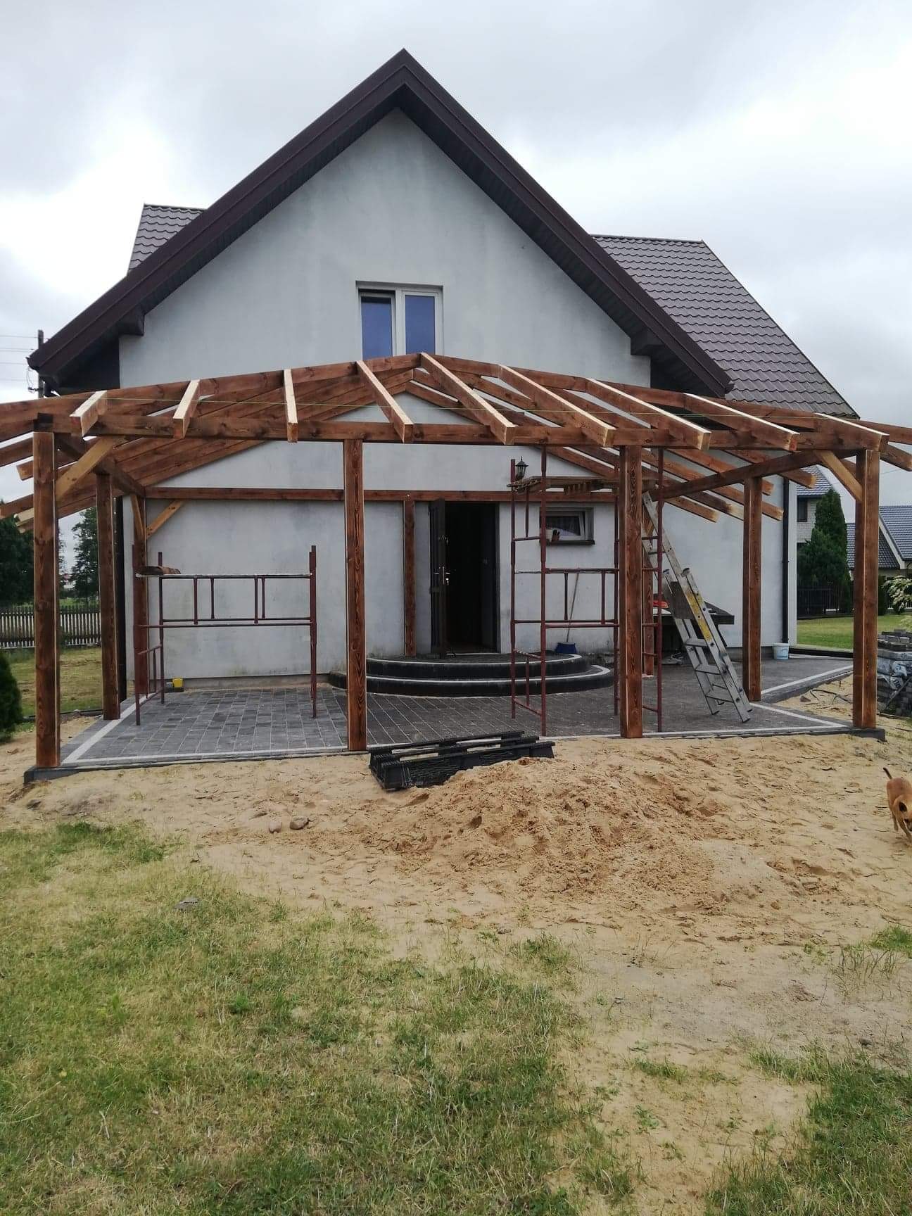 Drewniana konstrukcja z belek, prawdopodobnie zadaszenie tarasu, w trakcie budowy przed domem jednorodzinnym, widoczne rusztowania i piasek.
