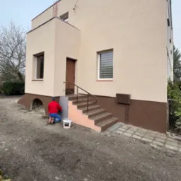 Pracownik w czerwonej kurtce i niebieskich spodniach, klęczący przed budynkiem z beżową elewacją, aplikuje szarą warstwę wykończeniową na cokół. Obok stoi białe wiadro z napisem BOLIX. Widoczny...