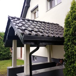 MichBud Usługi Budowlane - Zadaszenie nad wejściem do domu z ciemną dachówką, czarną rynną i jasnymi kolumnami. Obok rośnie wysoka tuja. Widoczna elewacja w jasnym odcieniu.
