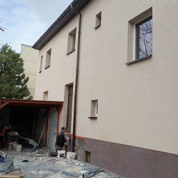 MichBud Usługi Budowlane - Pracownik nakłada tynk na elewację dwukondygnacyjnego domu z widoczną dolną, ciemniejszą partią. Na pierwszym planie wiadro z zaprawą i narzędzia.