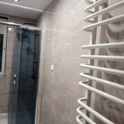 MichBud Usługi Budowlane - Wykończona łazienka z kabiną prysznicową i białym grzejnikiem drabinkowym na ścianie wyłożonej płytkami imitującymi kamień w odcieniach szarości i beżu.
