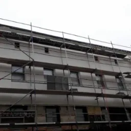 Budynek w Piotrków Kujawski w trakcie prac elewacyjnych, widoczne rusztowania, okna zabezpieczone folią, częściowo otynkowana ściana z widocznymi napisami.