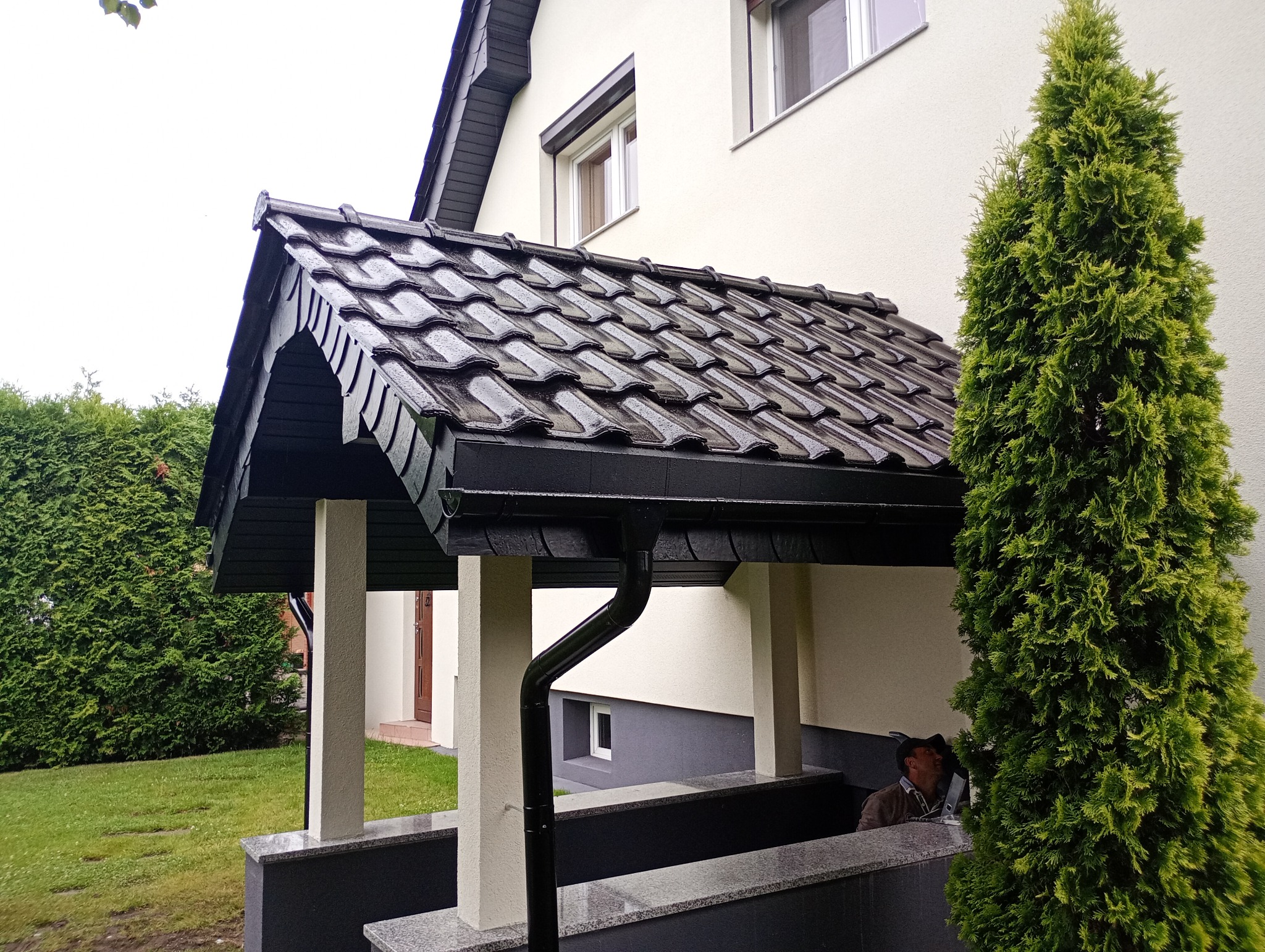 Zadaszenie nad wejściem do domu z ciemną dachówką, czarną rynną i jasnymi kolumnami. Obok rośnie wysoka tuja. Widoczna elewacja w jasnym odcieniu.