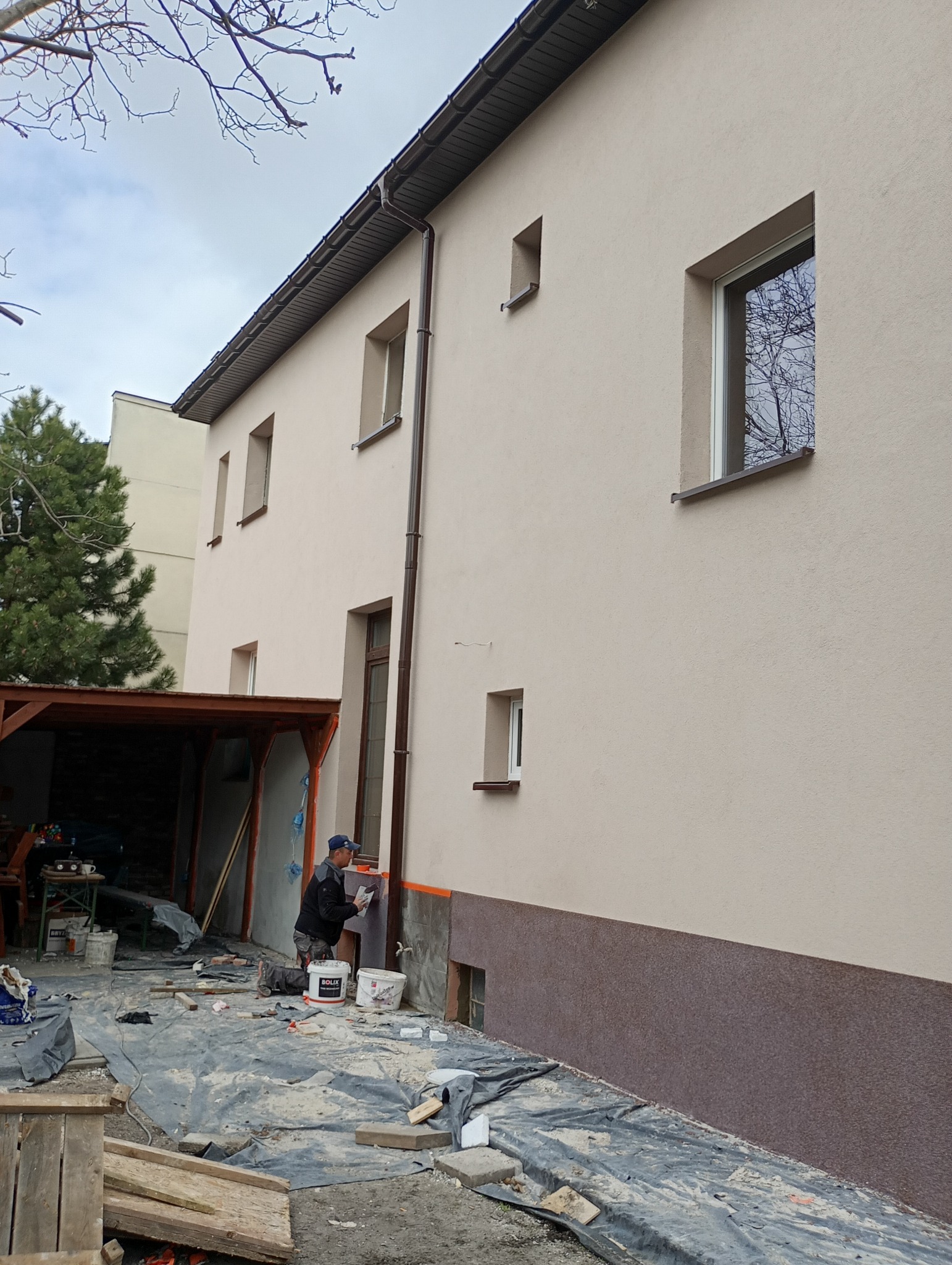 Pracownik nakłada tynk na elewację dwukondygnacyjnego domu z widoczną dolną, ciemniejszą partią. Na pierwszym planie wiadro z zaprawą i narzędzia.