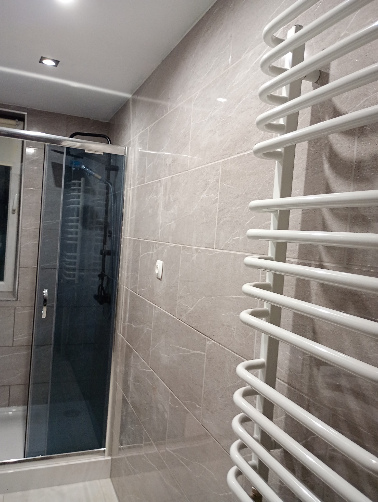 Wykończona łazienka z kabiną prysznicową i białym grzejnikiem drabinkowym na ścianie wyłożonej płytkami imitującymi kamień w odcieniach szarości i beżu.