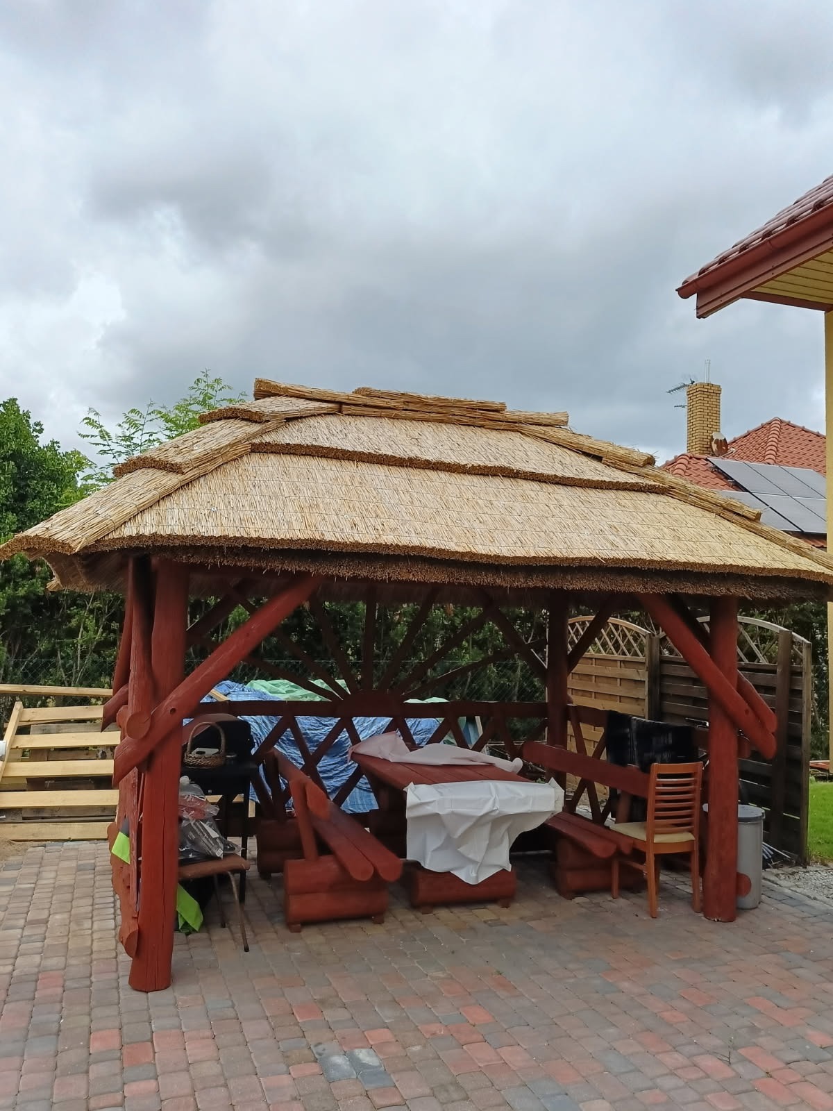 Kryty strzechą, drewniany altanowy dach w rustykalnym stylu. Czerwone, impregnowane drewno konstrukcji. W tle dom z panelami słonecznymi i ceglanym kominem.