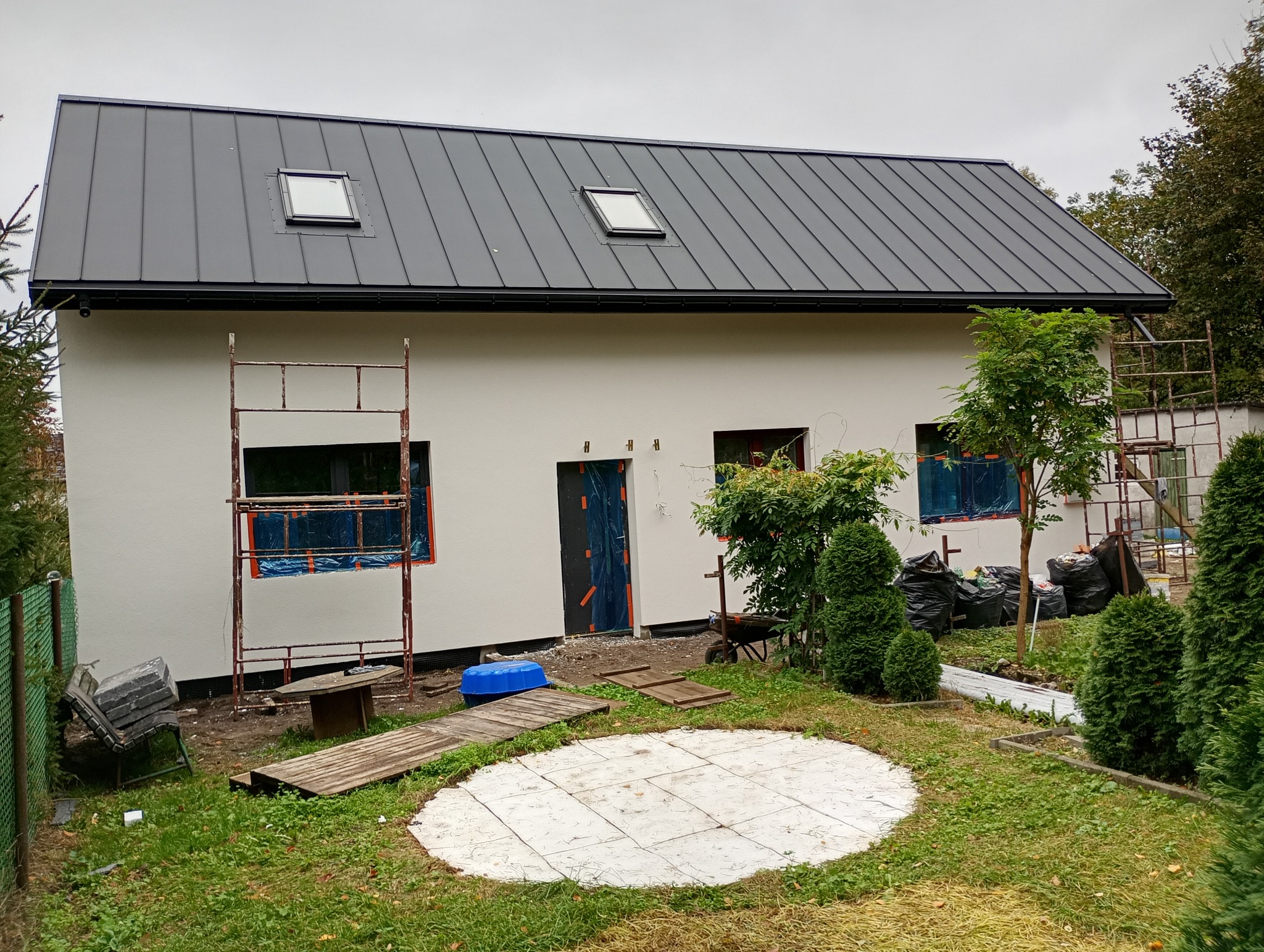 Dom jednorodzinny z szarą blachą na dachu i białą elewacją. Okna i drzwi zabezpieczone folią, obok rusztowania. Widoczne elementy ogrodu i przygotowania do dalszych prac.