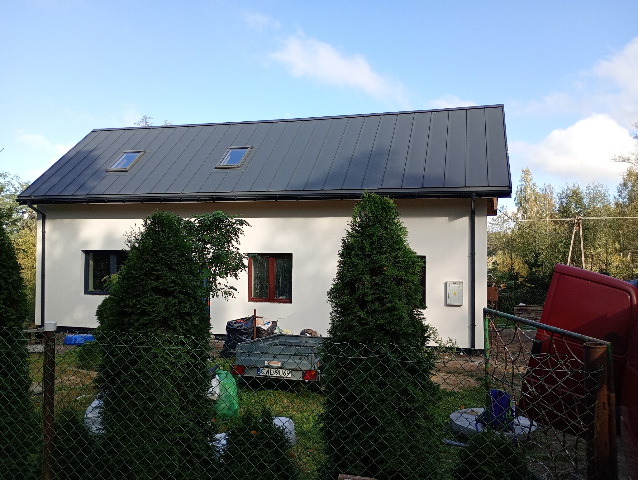 Biały dom jednorodzinny z szarym dachem blaszanym i oknami dachowymi, widoczny zza ogrodzenia i drzew iglastych. W tle zieleń drzew i niebieskie niebo.