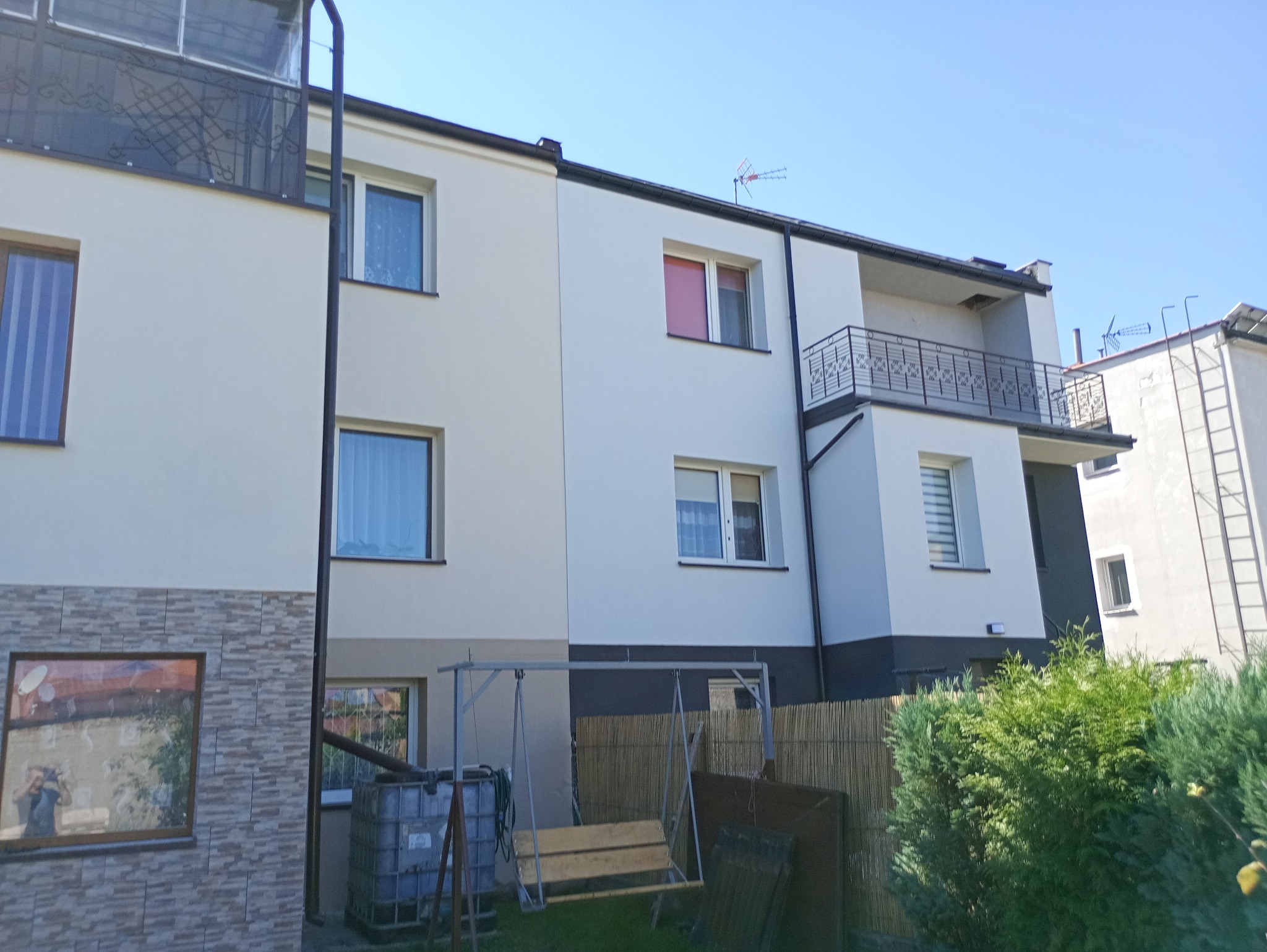 Elewacja dwukondygnacyjnego budynku z balkonem, pomalowana na biało z ciemnoszarym pasem na dole. Widoczne okna z roletami i anteną na dachu sąsiedniego budynku.