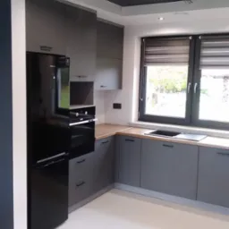 Nowoczesna, szara kuchnia z drewnianym blatem, czarną lodówką Samsung i oknem z roletami dzień-noc. Widoczne punktowe oświetlenie sufitowe i biała podłoga z płytek.