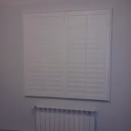 Białe okiennice wewnętrzne zamontowane w oknie, widoczny biały grzejnik pod oknem.