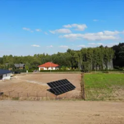 Czarny panel fotowoltaiczny na pustym polu, w tle domy z czerwonymi i ciemnymi dachami oraz las pod błękitnym niebem.