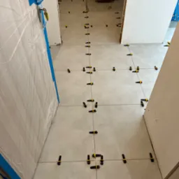 Układanie jasnych płytek podłogowych z użyciem systemu poziomowania, widoczne czarne klipsy i żółte kliny, zabezpieczone drzwi folią malarską z niebieską taśmą.