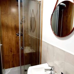 Adrian Malik Usługi Wykończeniowe - Nowoczesna kabina prysznicowa z drewnopodobnymi płytkami, okrągłe lustro z brązowym paskiem, biała umywalka z szafką i otwartą szufladą z zapasem papieru toaletowego.