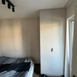 Neutralna sypialnia z łóżkiem w szaro-czarną pościel, gładkimi ścianami w odcieniach beżu i wysoką, białą szafą z minimalistycznymi, czarnymi uchwytami, oświetlona trzema punktowymi lampami...