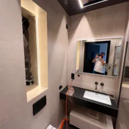 Nowoczesna łazienka z podświetlaną wnęką, czarnym blatem i lustrem, w którym odbija się osoba robiąca zdjęcie. Widoczna toaleta i elementy wykończenia w beżowych tonacjach.