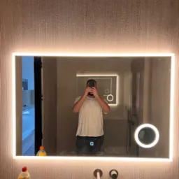 Nowoczesne lustro LED w łazience z fakturą na ścianie. Widoczne czarne baterie umywalkowe i odbicie wnętrza łazienki. Nietypowe oświetlenie tworzy przyjemny klimat.