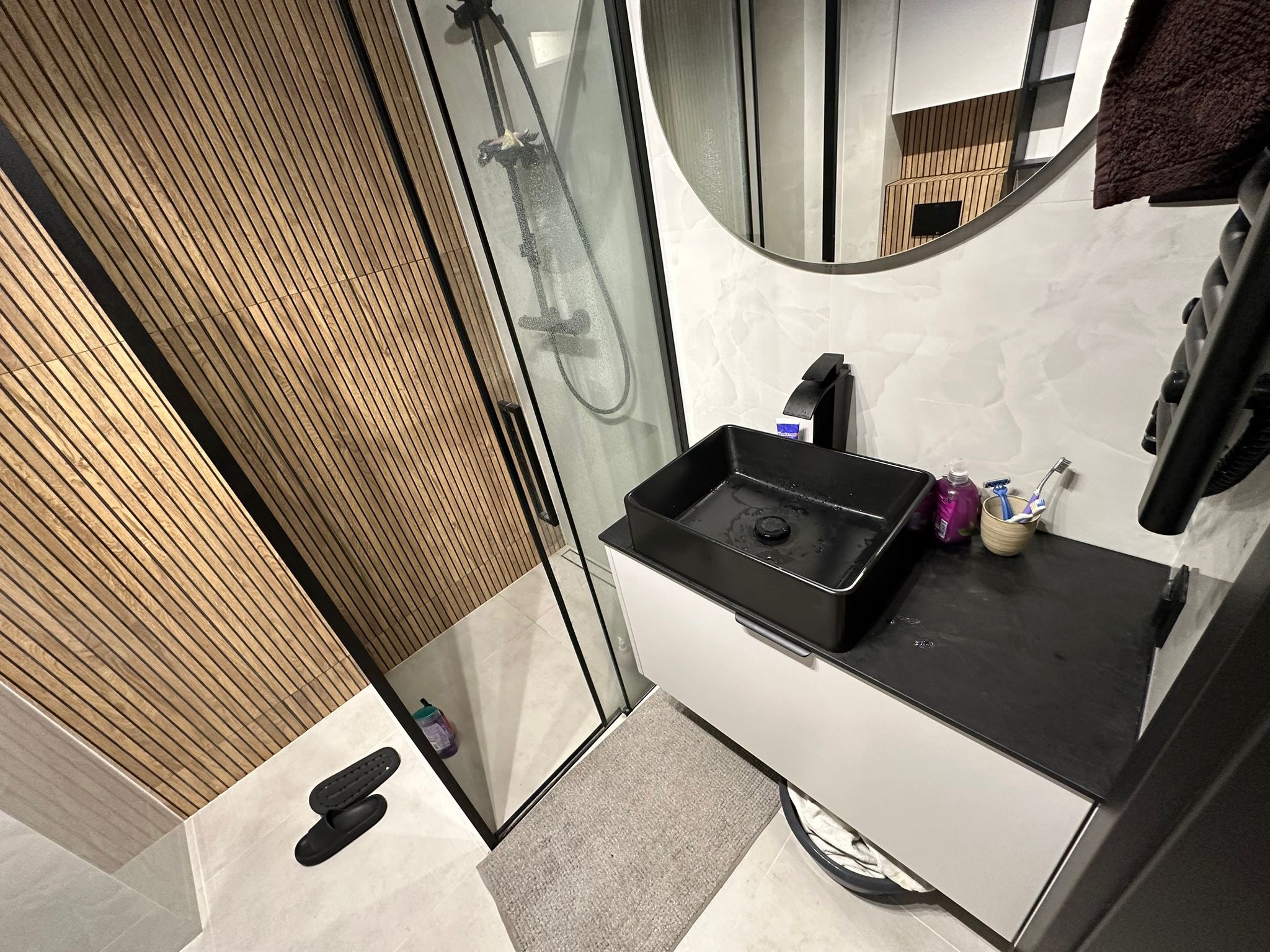 Nowoczesna łazienka z drewnianymi panelami ściennymi, czarną armaturą, kabiną prysznicową z czarnymi ramami i okrągłym lustrem.