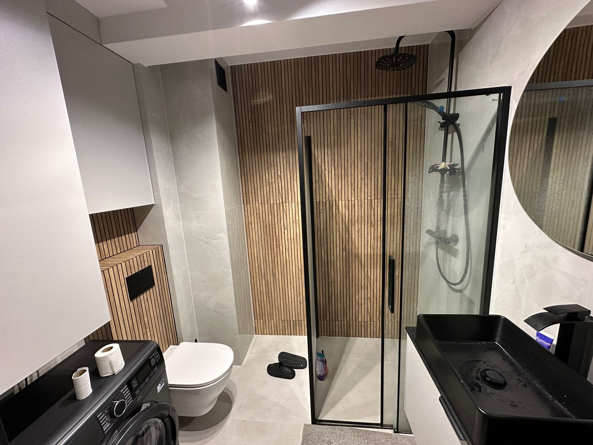 Nowoczesna łazienka z kabiną prysznicową typu walk-in, drewnianą ścianą dekoracyjną, czarną armaturą i pralką z rolkami papieru toaletowego na górze.