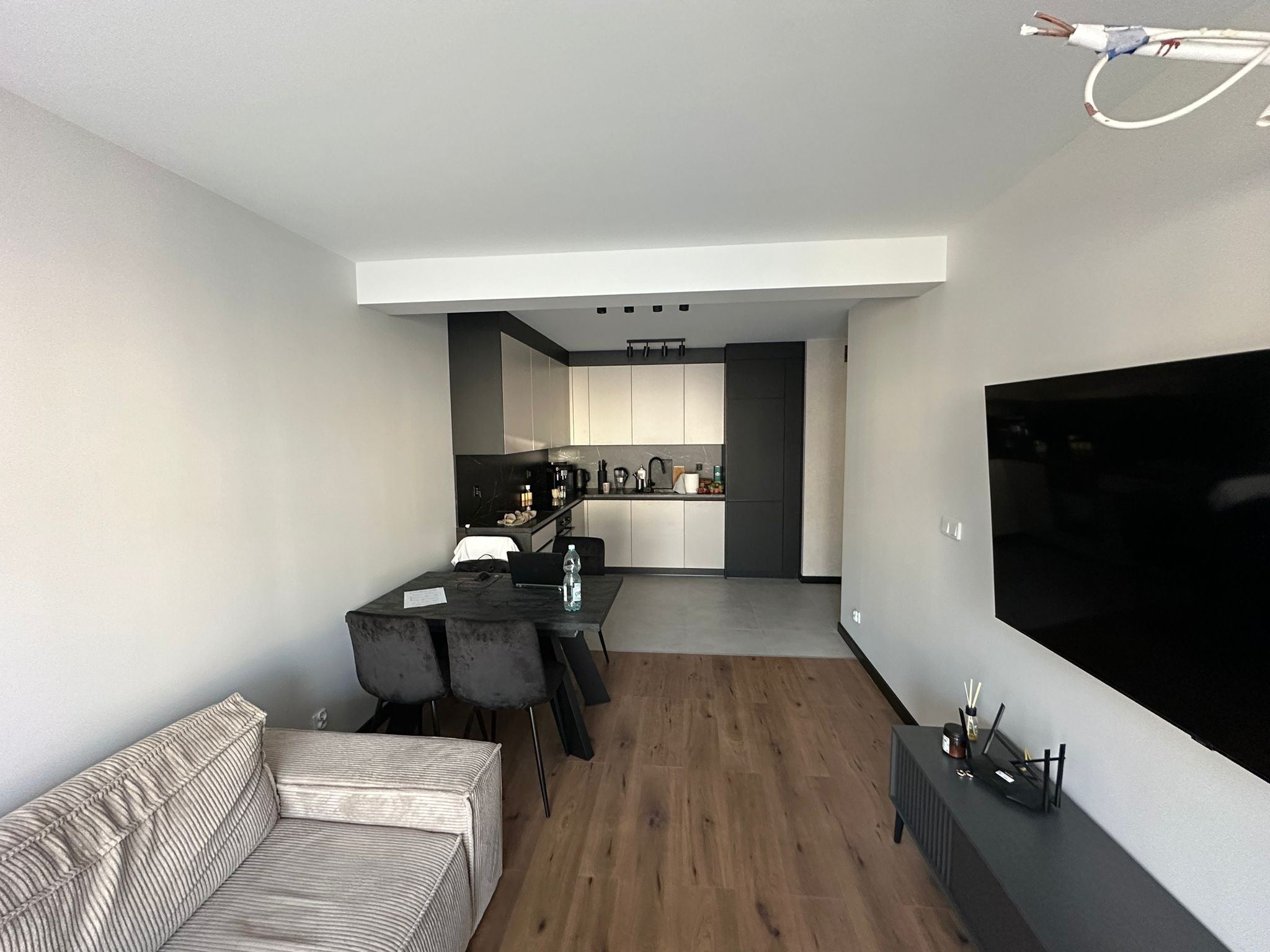 Nowoczesne wnętrze mieszkania z otwartą kuchnią, drewnianą podłogą i szarą sofą, widoczne wystające przewody elektryczne w rogu sufitu.