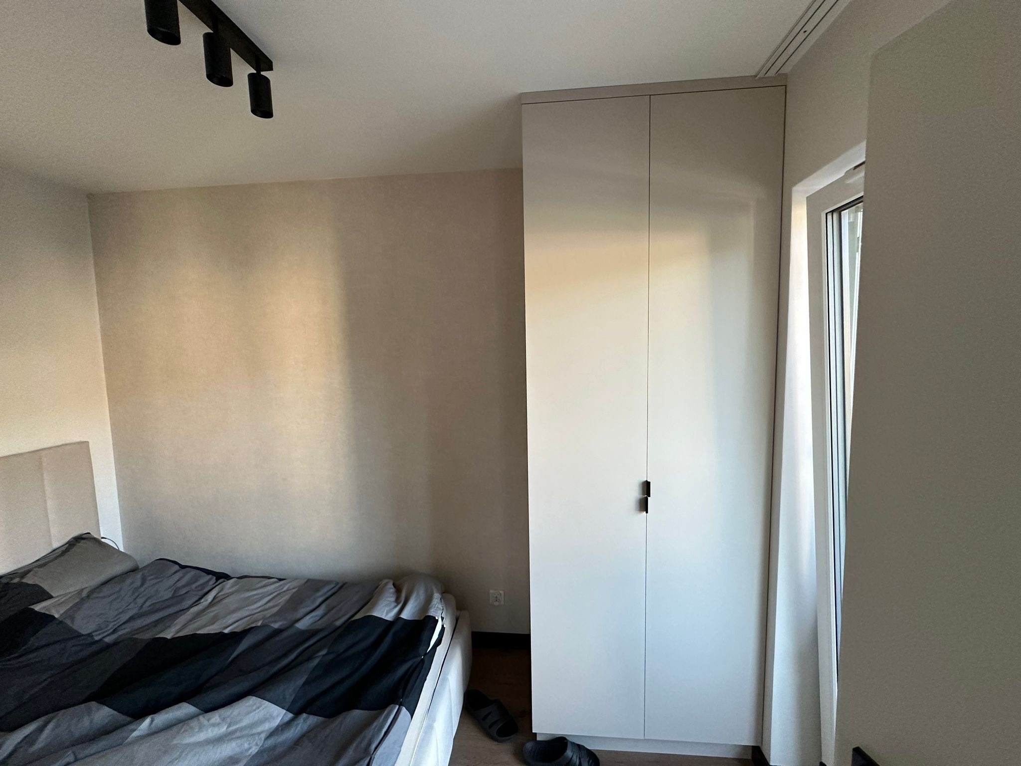 Neutralna sypialnia z łóżkiem w szaro-czarną pościel, gładkimi ścianami w odcieniach beżu i wysoką, białą szafą z minimalistycznymi, czarnymi uchwytami, oświetlona trzema punktowymi lampami...