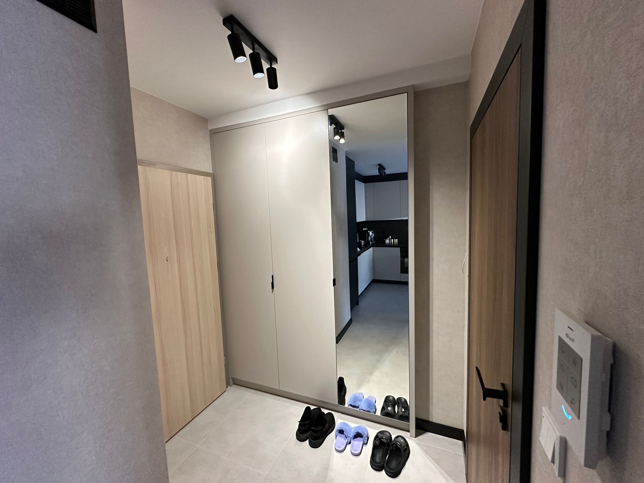 Przedpokój z przesuwną szafą z lustrem, w której odbija się kuchnia, na podłodze leżą kapcie, a na ścianie wisi panel sterowania.