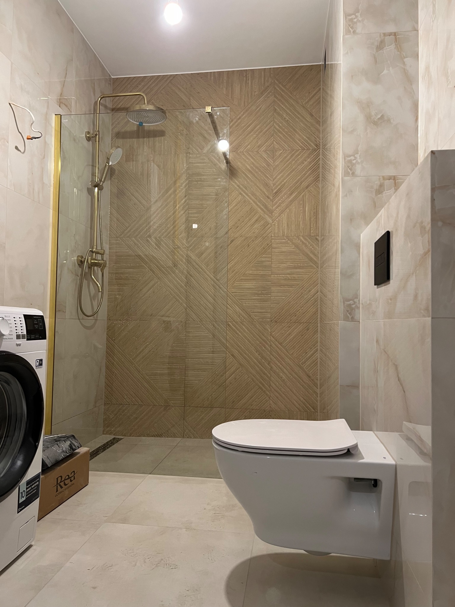 Nowoczesna łazienka po renowacji z kabiną prysznicową ze złotą armaturą, wiszącą toaletą i pralką Electrolux. Na podłodze i ścianach jasne płytki imitujące marmur, na ścianie pod prysznicem płytki...