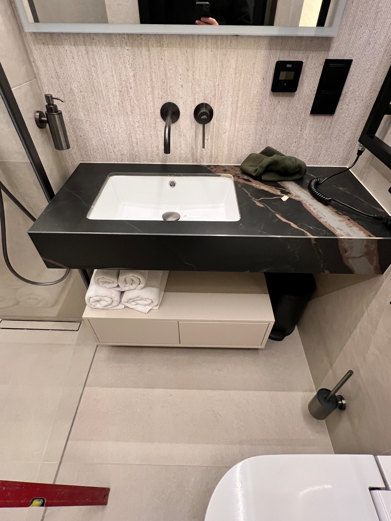 Nowoczesna łazienka: biała umywalka z czarnym blatem imitującym marmur, czarne baterie, beżowe płytki i białe ręczniki. Minimalistyczny design z akcentami kolorystycznymi.