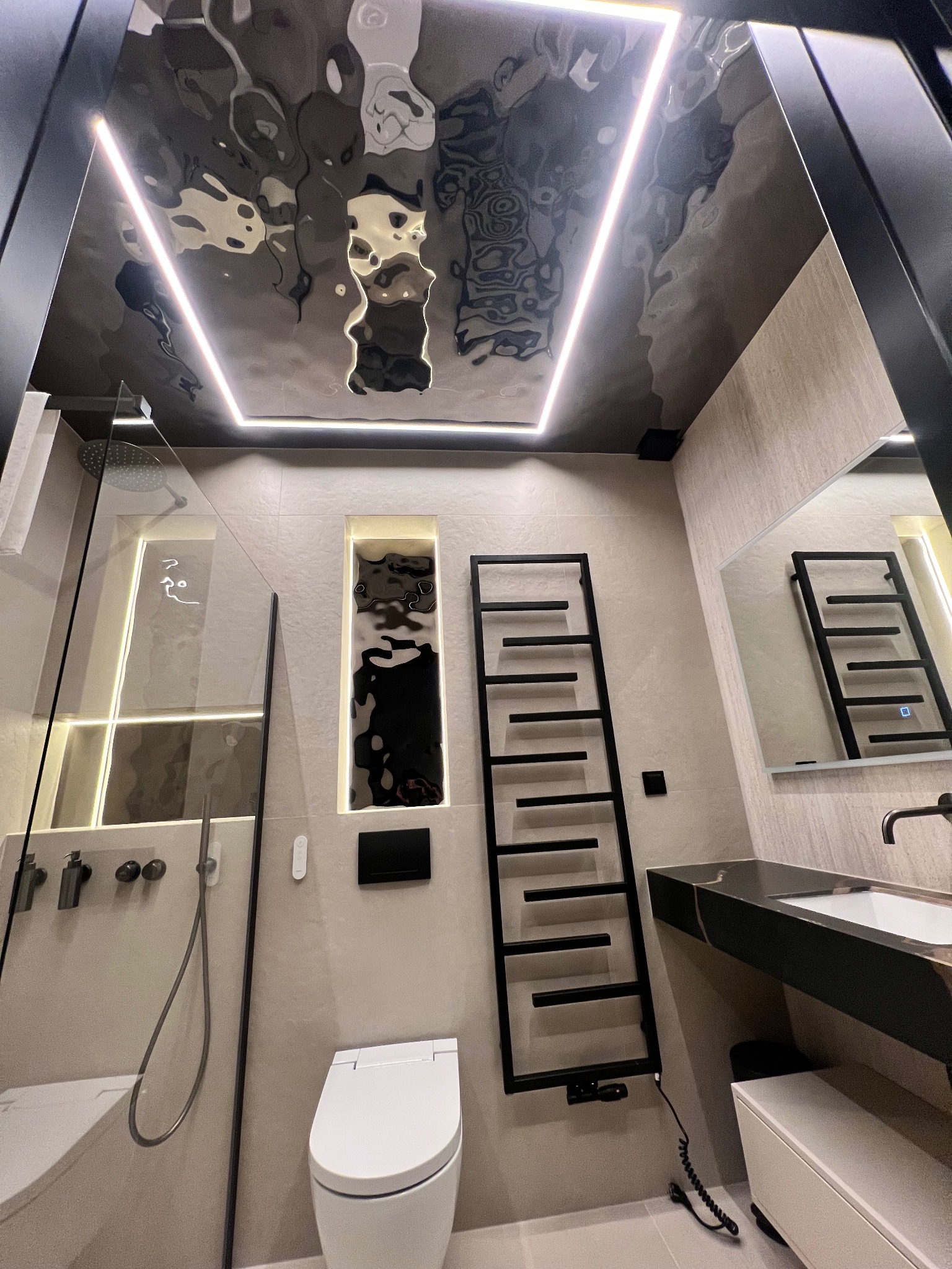 Nowoczesna łazienka z lustrzanym sufitem i oświetleniem LED, kabiną prysznicową ze szkła, grzejnikiem drabinkowym i umywalką z czarnym blatem.