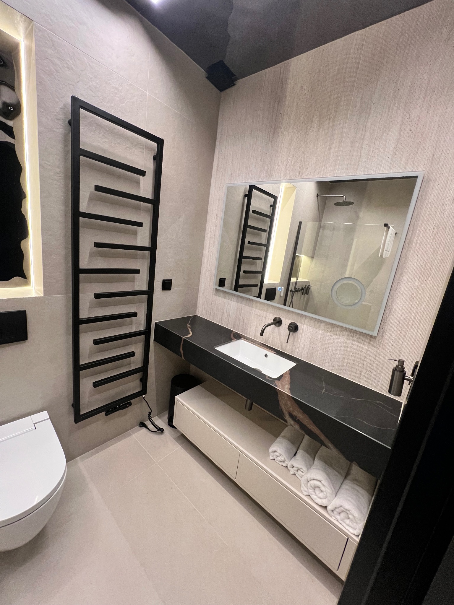 Nowoczesna łazienka z czarnym grzejnikiem drabinkowym, marmurowym blatem i podświetlanym lustrem. Minimalistyczny design, beżowe płytki, biała ceramika.