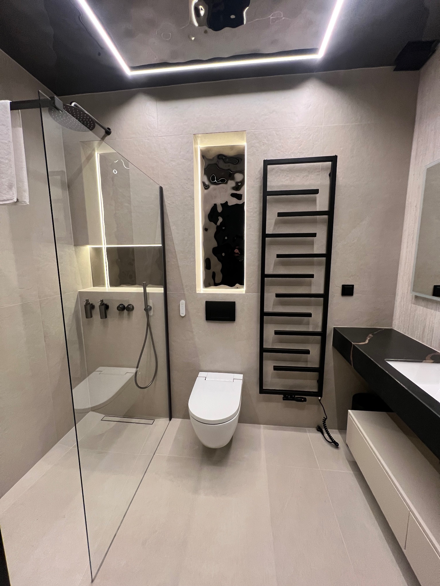 Nowoczesna łazienka z kabiną prysznicową typu walk-in, grzejnikiem drabinkowym i podświetlaną wnęką w Tylmanowej. Minimalistyczny design, jasne płytki, czarne akcenty.